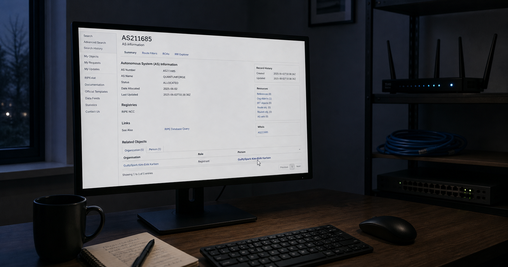 A dimly lit home office with a monitor displaying the RIPE database entry for AS211685, cursor hovering over the registrant name, an unplugged network cable on the desk.