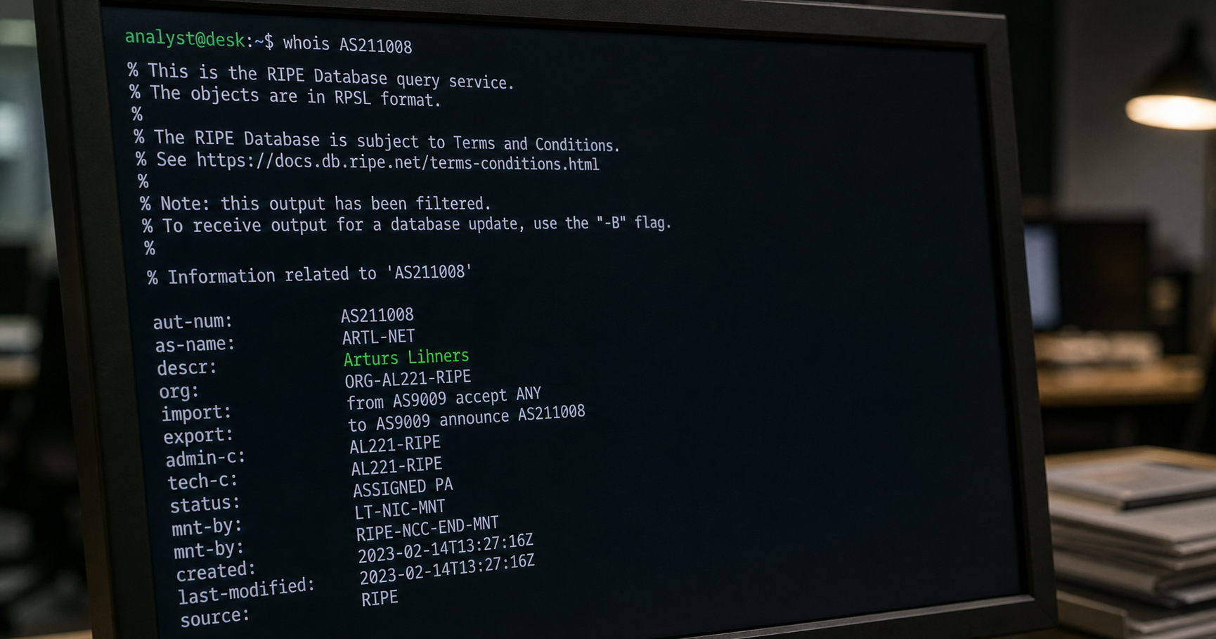 A computer terminal in a dim network operations center displaying a WHOIS query for AS211008 with the name 'Arturs Lihners' highlighted.